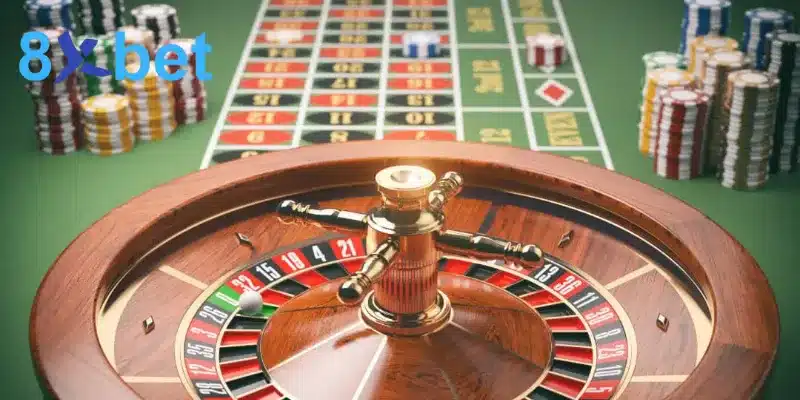 Vòng quay roulette có bi trắng phía trước bảng cược màu xanh và các chồng chip