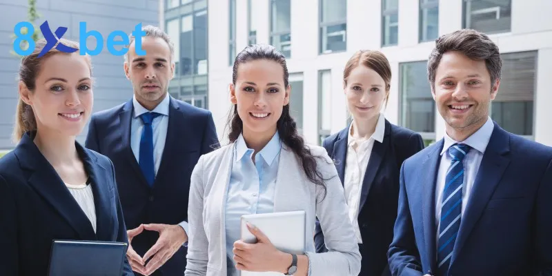 Năm nhân sự mặc vest đứng cười trước tòa nhà văn phòng